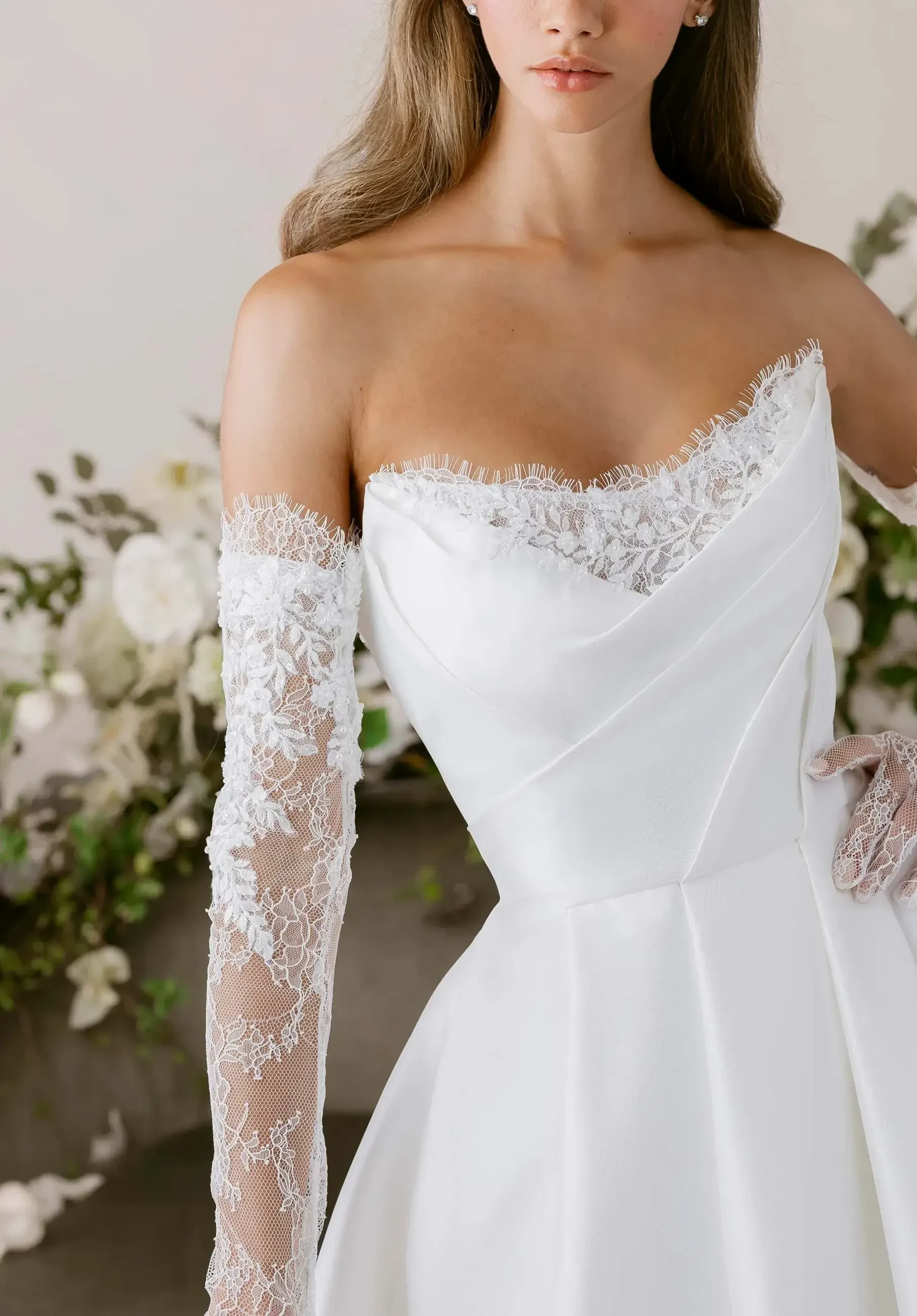 A close-up of a woman wearing an elegant off-the-shoulder white gown with intricate lace detail on the long sleeves, set against a floral background.