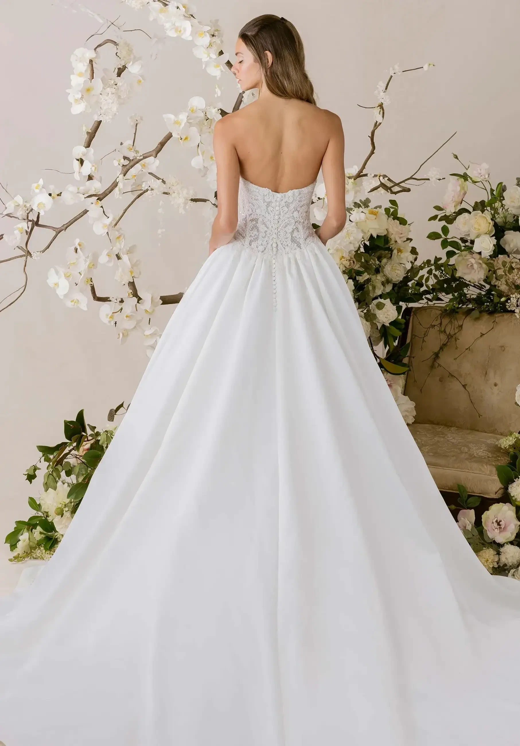 A bride in a strapless, embroidered white gown with a long train stands before lush white flowers and branches, creating a serene, elegant scene.