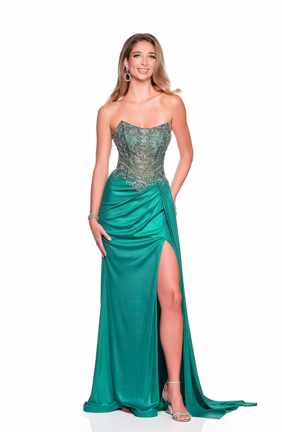 A woman smiles in a strapless emerald gown with beaded bodice, side slit, and train. She wears earrings and heels, exuding elegance and confidence.