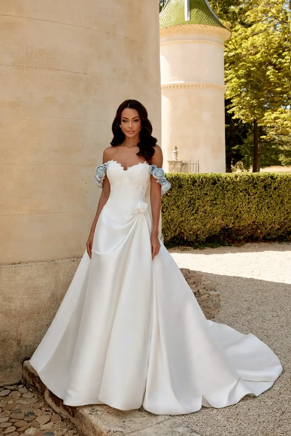 A woman in a white off-the-shoulder wedding dress with flutter sleeves, standing outside near a stone wall and greenery.