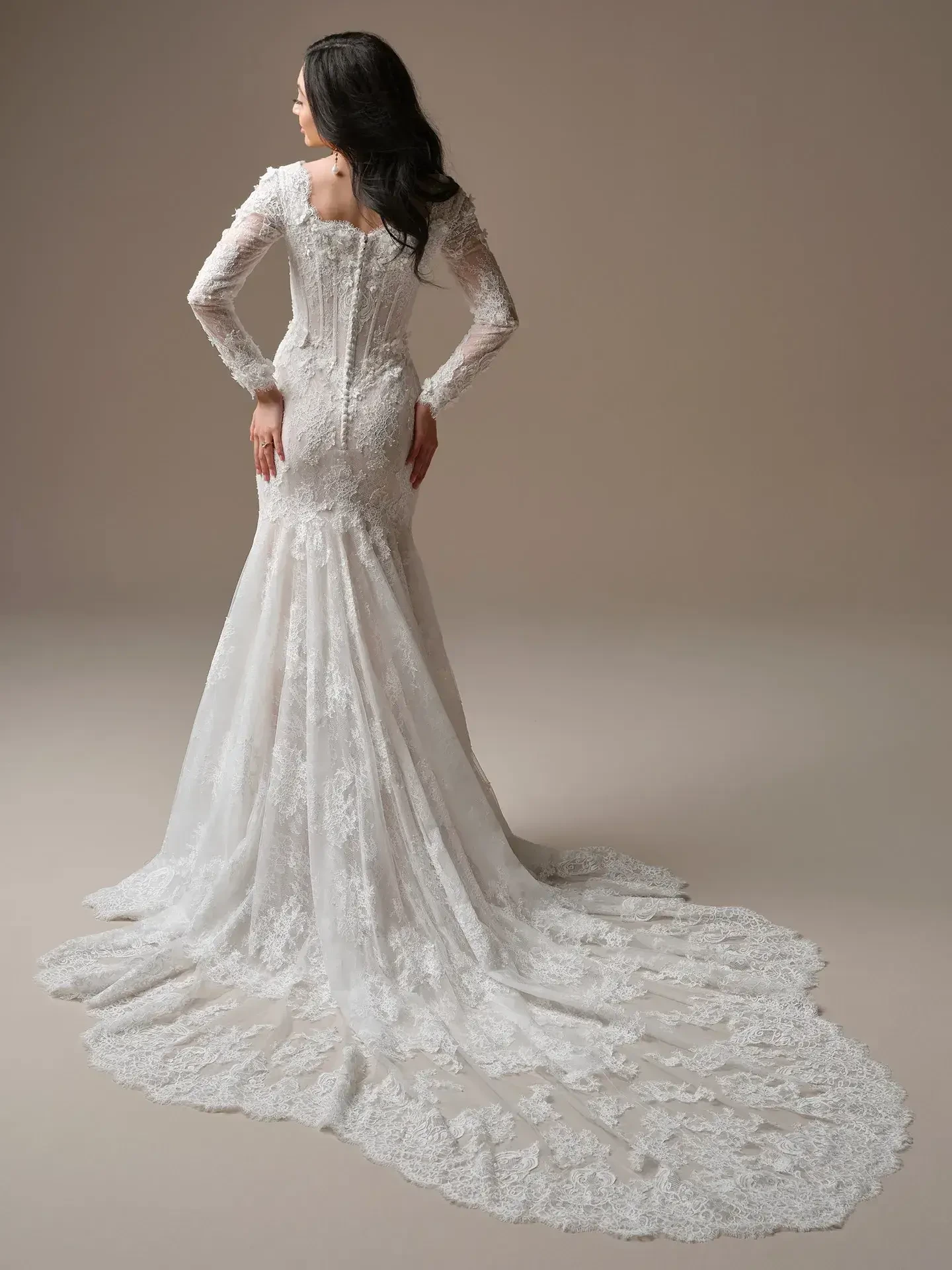 A woman in a long-sleeved lace wedding gown stands with her back facing, hands on hips. The elegant train flows gracefully, set against a neutral backdrop.