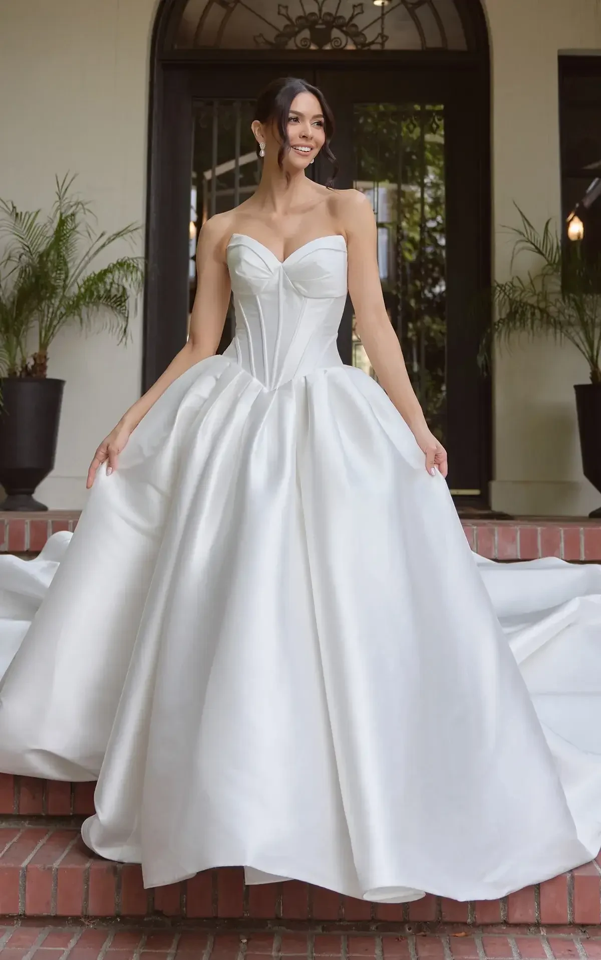 A smiling woman in a white wedding dress is standing on steps outside a building, with plants in the background.