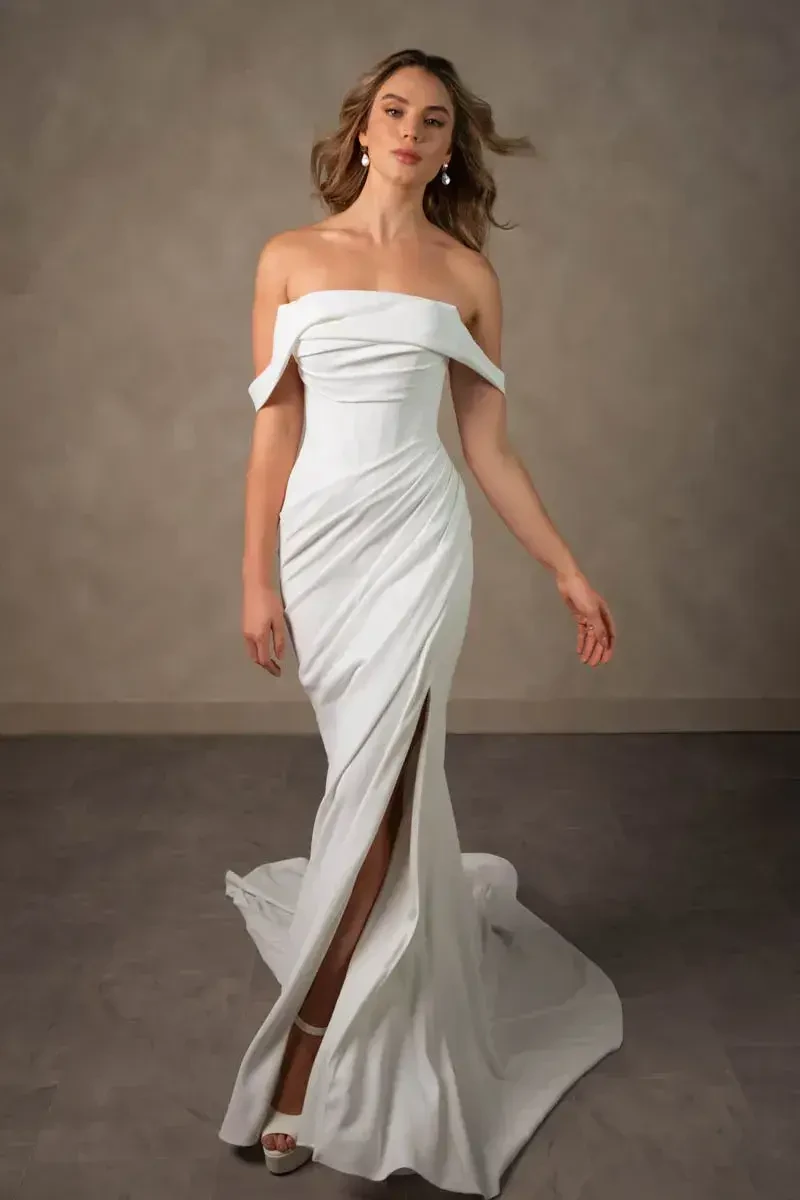 A model wearing an elegant off-shoulder white dress with a front slit, standing against a neutral background.