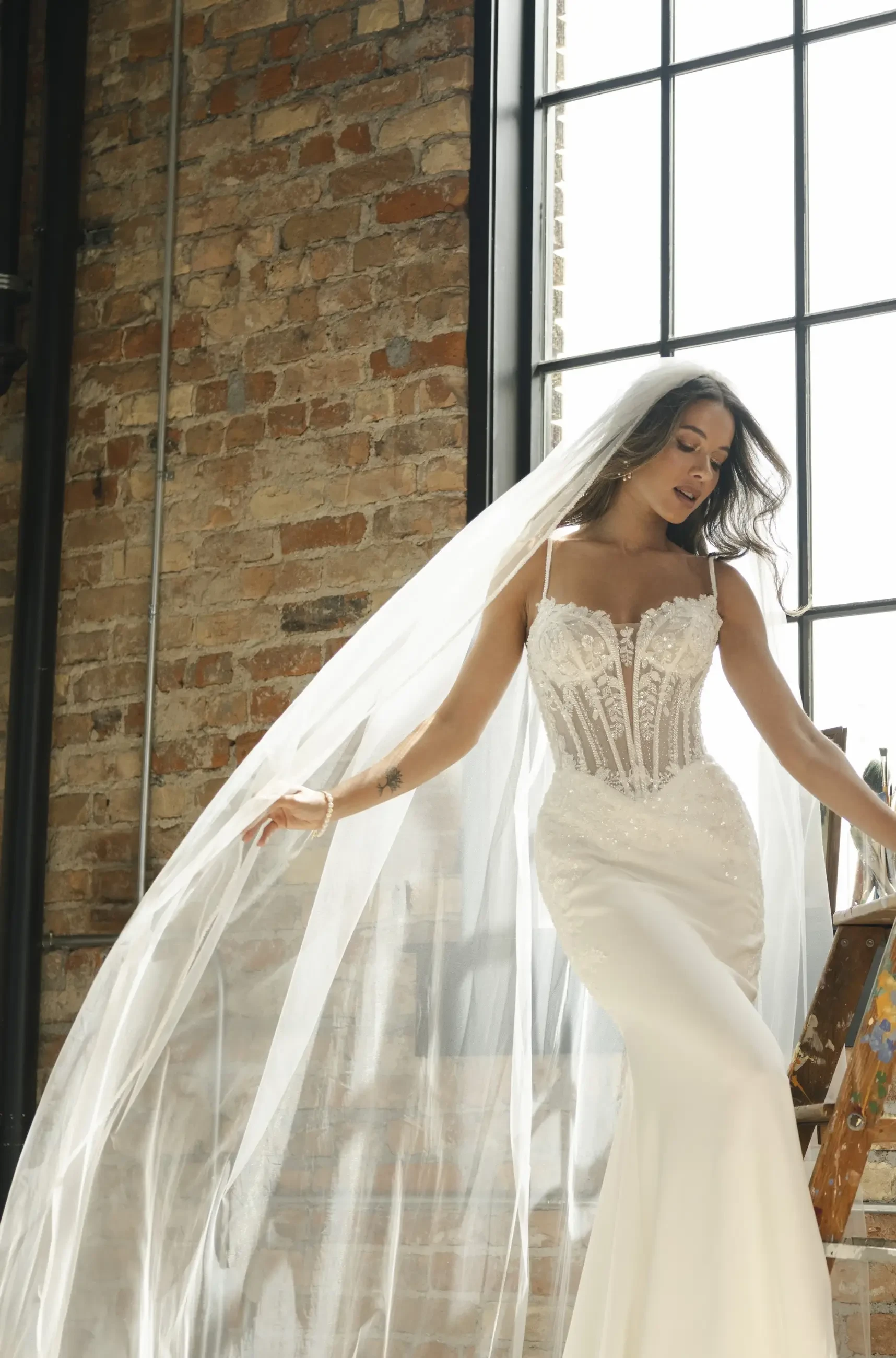 A woman in a bridal gown with a lace bodice and flowing veil, posing in front of a large window with brick walls in the background.