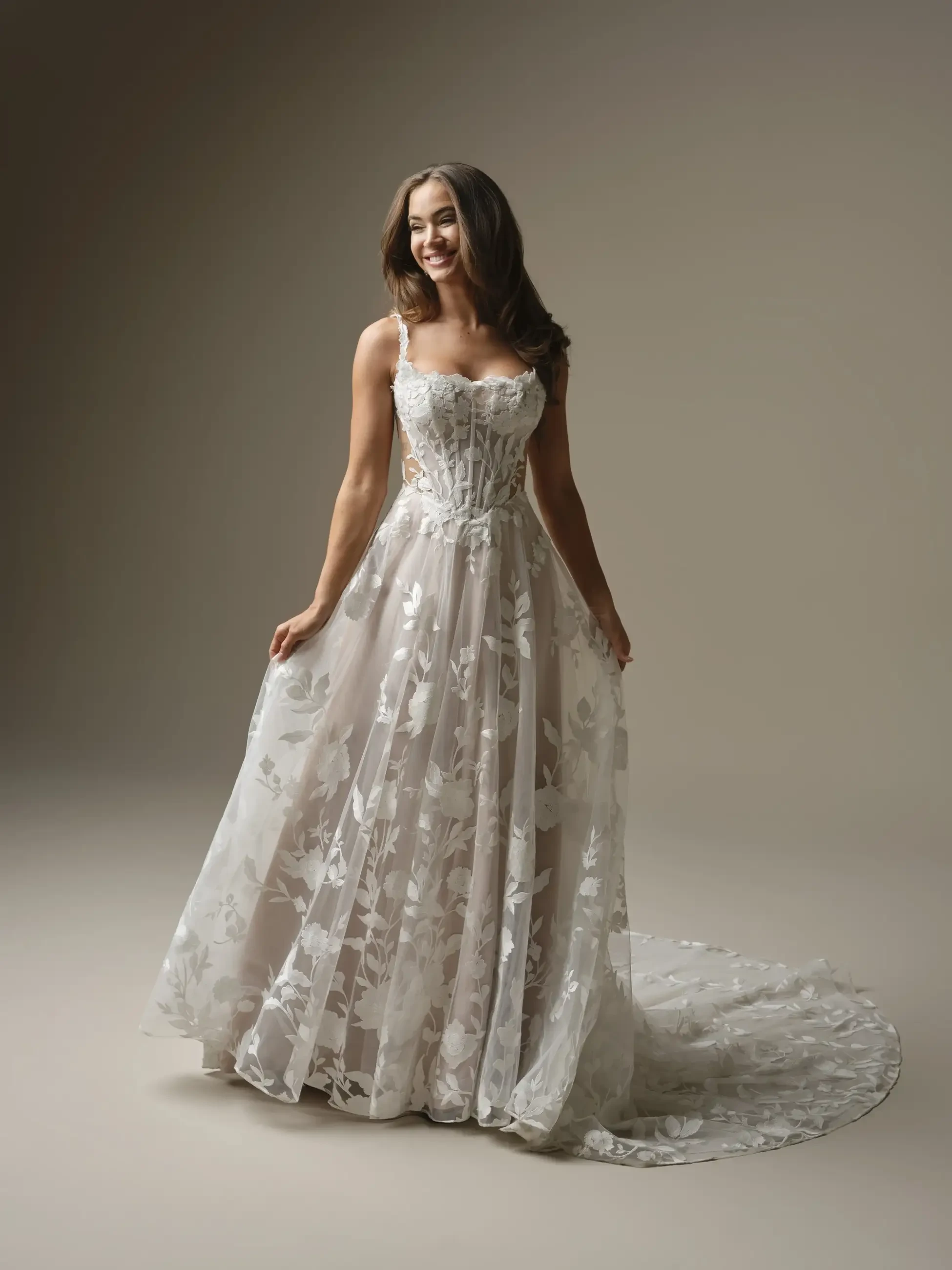 A woman wearing a beautiful, elegant gown with floral patterns and a flowing train, standing in front of a neutral background.