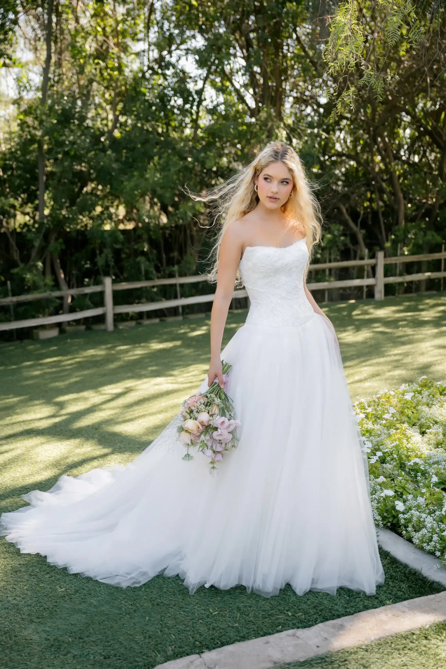Bride in strapless white gown holding a bouquet stands on grass, surrounded by trees, exuding elegance and tranquility under soft sunlight.