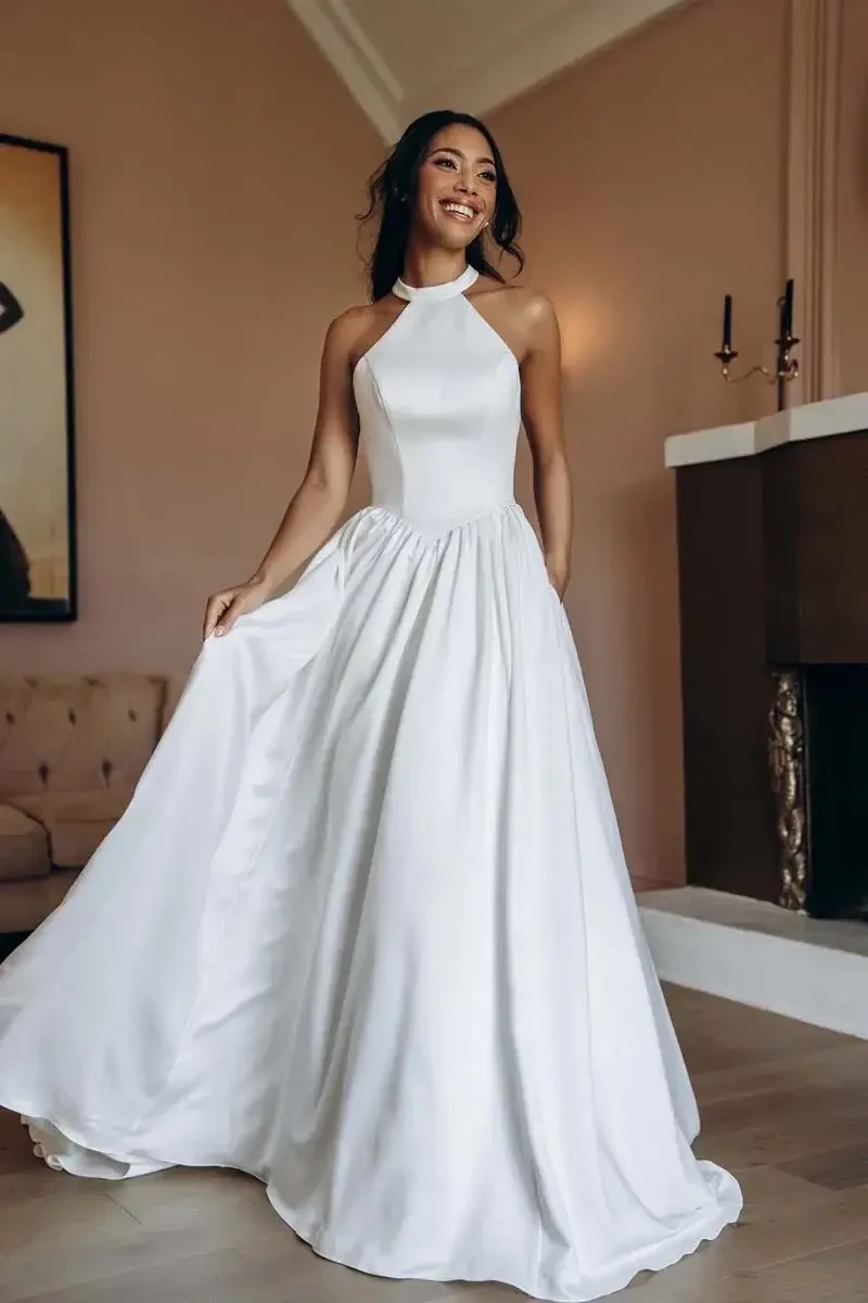 A woman wearing a white halter neck dress, standing and holding the skirt of her dress, with a stylish interior in the background.